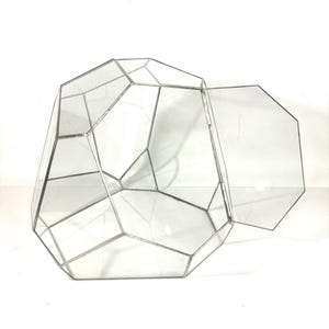 Peut inclure: Terrarium géométrique en verre avec couvercle à charnière. Les panneaux de verre transparents sont encadrés de métal argenté, créant un design angulaire moderne. Le terrarium est ouvert, révélant l'espace intérieur, idéal pour les plantes.