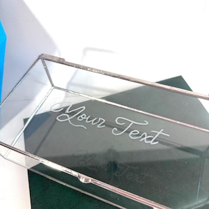 May include: A clear glass rectangular box with silver trim. The words "Your Text" are etched on the glass. The box is resting on a dark green surface.