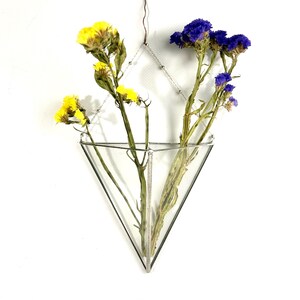 以下が含まれることがあります： 銀色のチェーンで吊るされた、三角形のクリアなガラスの花瓶。黄色と青色のドライフラワーが入っています。花瓶は白い背景に吊るされています。