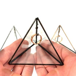 Puede incluir: Un portaanillos geométrico de vidrio y metal con forma de pirámide. El anillo es dorado y está suspendido dentro de la pirámide. El marco de metal es de color marrón oscuro. Se ven otros dos portaanillos en el fondo.