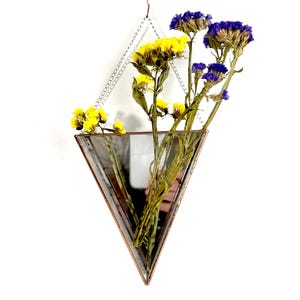 Może przedstawiać: Szklany wazon w kształcie trójkąta z lustrzanym wnętrzem, wiszący na srebrnym łańcuchu. Wazon zawiera suszone żółte i fioletowe kwiaty.