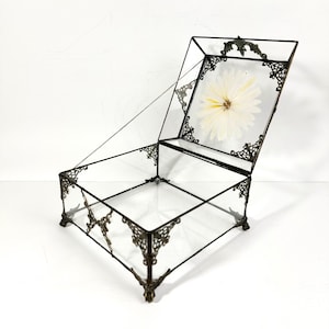 May include: An open, clear glass box with a hinged lid, featuring ornate bronze-colored metal detailing. The lid displays a preserved white flower. The box is designed with decorative corner feet and a chain to hold the lid open.