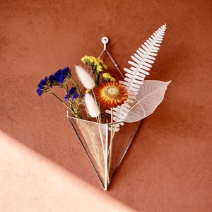 May include: A wall-mounted glass vase in a triangular shape, filled with dried flowers and foliage. The arrangement includes blue, yellow, and orange blooms, along with white fluffy accents and a white leaf. The vase hangs from a chain against a reddish-brown wall.