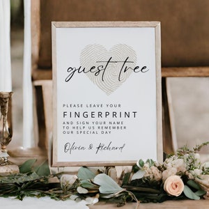 Può includere: Un cartello bianco con cornice in legno marrone, che presenta un design a forma di cuore con un'impronta digitale e il testo "guest tree". Il cartello invita gli ospiti a lasciare la loro impronta digitale e firmare il loro nome per aiutare a ricordare il giorno speciale di Olivia e Richard.