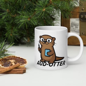 May include: White ceramic mug featuring a cartoon otter wearing glasses and holding a pen and notepad. The text "AUD-OTTER" is printed below the otter. The mug has a white handle and a white interior.