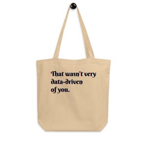 Puede incluir: Bolso tote beige con texto negro "That wasn't very data-driven of you". El bolso tiene asas largas y un diseño sencillo, adecuado para llevar objetos. El bolso está hecho de una tela suave de aspecto natural, ideal para el uso diario.