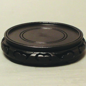 May include: A black wooden pedestal with a carved design around the edge. The pedestal is round and has a smooth, polished surface.