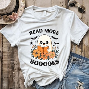 以下が含まれることがあります： 本を読む漫画の幽霊が描かれた白いTシャツ。オレンジ色の花、コウモリ、クモに囲まれています。Tシャツには黒で「READ MORE BOOOOKS」と書かれています。読書好きのための遊び心のあるデザインです。
