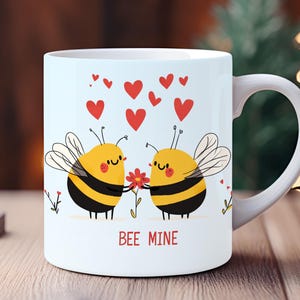 May include: White ceramic mug featuring a cartoon design of two smiling bees holding a flower, with red hearts above them. The text "BEE MINE" is printed below the bees. The mug has a white handle and is set on a wooden surface.