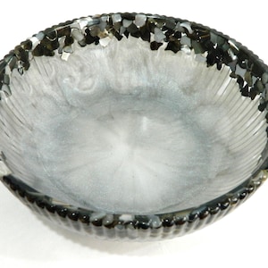 Puede incluir: Un cuenco decorativo con un interior nacarado y un borde adornado con piedras oscuras de forma irregular. El interior del cuenco tiene un efecto arremolinado e iridiscente, creando una estética única y elegante. El borde tiene un diseño texturizado y perlado.