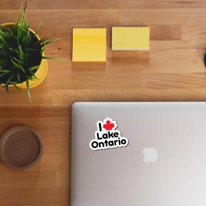May include: A die-cut vinyl sticker featuring the text I Lake Ontario in black sans-serif font, with a red Canadian maple leaf replacing the heart symbol. The sticker has a white border and is placed on the lid of a silver laptop computer.