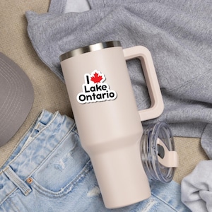May include: A light pink insulated tumbler with a handle and a clear lid. The tumbler features a sticker that reads I, followed by a red maple leaf, and the text Lake Ontario in black letters. The tumbler has a metallic rim at the top.