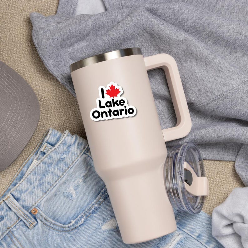May include: A light pink insulated tumbler with a handle and a clear lid. The tumbler features a sticker that reads I, followed by a red maple leaf, and the text Lake Ontario in black letters. The tumbler has a metallic rim at the top.