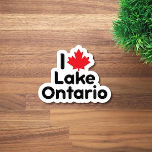 May include: A white die-cut sticker featuring the black text I Lake Ontario with a red maple leaf symbol replacing the word heart. The sticker has a clean white border and is shaped to follow the contour of the bold, sans-serif lettering.