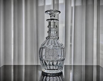 Pode incluir: Um decantador de vidro transparente com um padrão geométrico multifacetado, detalhes de corte de diamante no corpo e uma tampa de vidro a condizer. O recipiente tem uma base larga, um pescoço cónico e um topo circular.