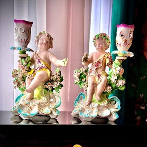 Puede incluir: Par de candelabros antiguos de porcelana. Cada candelabro presenta una figura de querubín adornada con guirnaldas florales y telas coloridas. Los querubines sostienen copas de velas florales. Las bases tienen diseños florales y de hojas, con detalles en azul y amarillo.