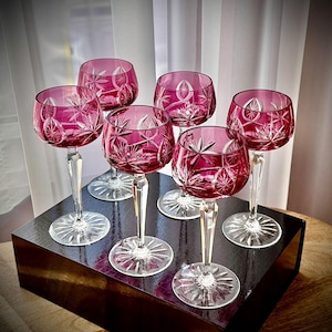 May include: A set of six cranberry-colored cut glass wine glasses featuring intricate starburst and geometric patterns on the bowls. Each glass has a clear, faceted stem and a circular base with a radial sunburst design.