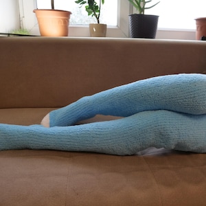 Könnte beinhalten: Ein Paar blaue, flauschige Beinstulpen mit weißem Besatz. Die Beinstulpen werden an den Beinen einer Person getragen und liegen auf einem braunen Sofa.