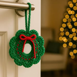 Puede incluir: Una corona de ganchillo verde hecha a mano con un lazo rojo y detalles de costura. La corona está colgada de un pomo de la puerta. Decoración festiva.