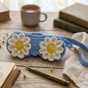 Puede incluir: Un estuche de gafas de ganchillo azul claro con dos grandes aplicaciones de margaritas. Las margaritas tienen pétalos color crema y centros amarillos. El estuche tiene una correa azul a juego con un cierre plateado. El estuche está sobre una superficie de madera.
