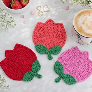 Könnte beinhalten: Drei handgefertigte Häkelteller in Tulpenform in Rot und Rosa mit grünen Blättern. Eine Tasse Kaffee mit Latte Art, eine Schüssel Erdbeeren und dekorative Gegenstände sind ebenfalls im Bild.