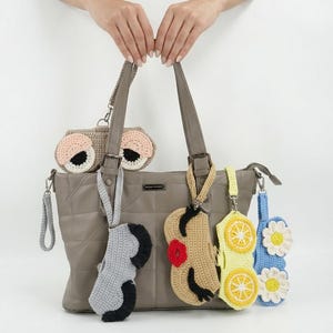 K&ouml;nnte beinhalten: Eine gesteppte Tragetasche in Taupe, an deren Riemen f&uuml;nf verschiedene H&auml;kelaccessoires h&auml;ngen. Die Accessoires zeigen verschiedene Designs, darunter Augen, ein Gesicht mit roten Lippen, gelbe Zitronenscheiben und wei&szlig;e G&auml;nsebl&uuml;mchen.