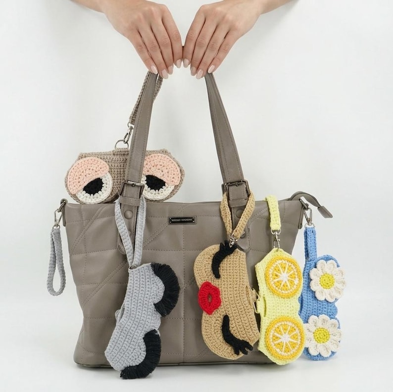K&ouml;nnte beinhalten: Eine gesteppte Tragetasche in Taupe, an deren Riemen f&uuml;nf verschiedene H&auml;kelaccessoires h&auml;ngen. Die Accessoires zeigen verschiedene Designs, darunter Augen, ein Gesicht mit roten Lippen, gelbe Zitronenscheiben und wei&szlig;e G&auml;nsebl&uuml;mchen.