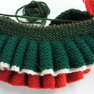 K&ouml;nnte beinhalten: Ein teilweise fertiggestellter Strickartikel in Weihnachtsfarben. Der Artikel wird von zwei H&auml;nden gehalten und zeigt das gr&uuml;ne, rote und wei&szlig;e Garn. Der Artikel hat einen ger&uuml;schten Rand und ein geripptes Oberteil.
