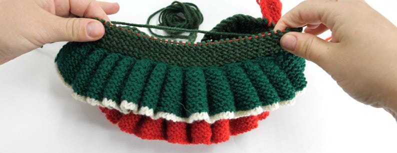 K&ouml;nnte beinhalten: Ein teilweise fertiggestellter Strickartikel in Weihnachtsfarben. Der Artikel wird von zwei H&auml;nden gehalten und zeigt das gr&uuml;ne, rote und wei&szlig;e Garn. Der Artikel hat einen ger&uuml;schten Rand und ein geripptes Oberteil.