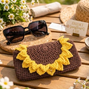 Op de afbeelding: Een bruine gehaakte etui met een gele zonnebloem, zonnebril en strohoed op een houten oppervlak. De etui is rechthoekig en heeft een gehaakte zonnebloemrand. Een label leest "CROCHET SUNFLOWER CASE."
