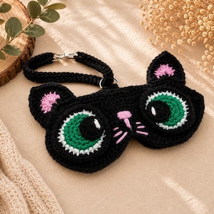 May include: A black crochet sleep mask shaped like a cat's face. The mask features large green eyes with white highlights, pink inner ears, and a pink nose and whiskers. It has a black strap with a silver clasp.