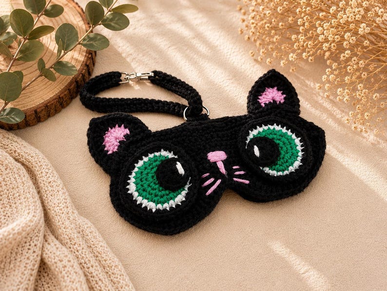 May include: A black crochet sleep mask shaped like a cat's face. The mask features large green eyes with white highlights, pink inner ears, and a pink nose and whiskers. It has a black strap with a silver clasp.