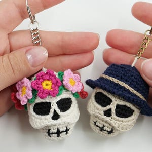 Könnte beinhalten: Zwei gehäkelte Totenkopf-Schlüsselanhänger. Ein Totenkopf ist cremeweiß mit schwarzen Augenhöhlen und Nase, verziert mit einer Krone aus rosa, magentafarbenen und gelben Blumen. Der andere Totenkopf ist cremeweiß mit schwarzen Details und trägt einen dunkelblauen Hut.