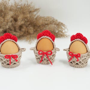 Puede incluir: Tres portahuevos de ganchillo hechos a mano. Cada soporte es beige con un lazo rojo y una parte superior roja con volantes. Los huevos son de color marrón claro. Los soportes están dispuestos en fila sobre una superficie blanca.