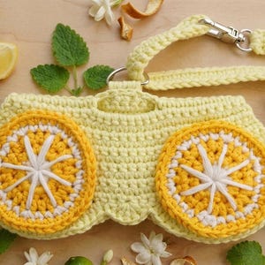 K&ouml;nnte beinhalten: Ein hellgelbes geh&auml;keltes T&auml;schchen mit zwei markanten kreisf&ouml;rmigen Zitronenscheiben-Motiven aus leuchtend gelbem und wei&szlig;em Garn. Das T&auml;schchen verf&uuml;gt &uuml;ber eine passende geh&auml;kelte Handschlaufe mit silberfarbenem Karabinerhaken.