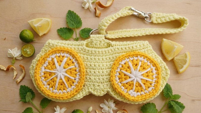 K&ouml;nnte beinhalten: Ein hellgelbes geh&auml;keltes T&auml;schchen mit zwei markanten kreisf&ouml;rmigen Zitronenscheiben-Motiven aus leuchtend gelbem und wei&szlig;em Garn. Das T&auml;schchen verf&uuml;gt &uuml;ber eine passende geh&auml;kelte Handschlaufe mit silberfarbenem Karabinerhaken.