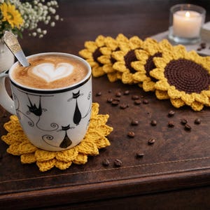 May include: A white mug with black cat illustrations, filled with coffee and a heart-shaped design in the foam, sits on a yellow crochet sunflower coaster. Crochet sunflower coasters and coffee beans are scattered on a wooden surface.