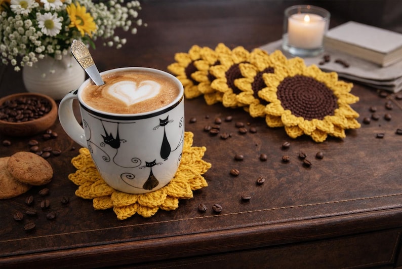 May include: A white mug with black cat illustrations, filled with coffee and a heart-shaped design in the foam, sits on a yellow crochet sunflower coaster. Crochet sunflower coasters and coffee beans are scattered on a wooden surface.