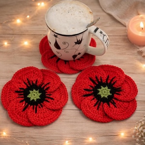 Puede incluir: Una taza blanca con diseño de gato descansa sobre un posavasos rojo de ganchillo. Se muestran dos posavasos de amapola roja de ganchillo adicionales. Una vela encendida y luces de hadas añaden un ambiente acogedor.