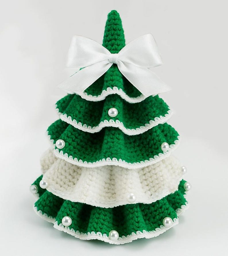 May include: A handmade, tiered Christmas tree decoration. The tree is crocheted with green and white yarn, adorned with white pearl beads and a white satin bow at the top. The tree is approximately 6 inches tall.
