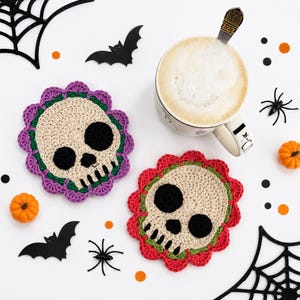 Könnte beinhalten: Zwei runde Häkeluntersetzer mit einem beigen Totenkopfdesign, schwarzen Augenhöhlen und Zähnen. Ein Untersetzer hat einen violetten, der andere einen roten, wellenförmigen Rand. Beide verfügen über einen dünnen grünen Ring um den Totenkopf.