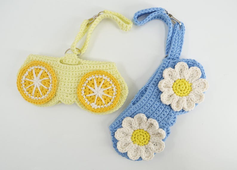 K&ouml;nnte beinhalten: Zwei geh&auml;kelte Taschen mit Handschlaufen. Eine Tasche ist hellgelb mit zwei orange-wei&szlig;en Zitrusfruchtmotiven. Die andere Tasche ist hellblau mit zwei wei&szlig;-gelben G&auml;nsebl&uuml;mchenmotiven. Beide Artikel verf&uuml;gen &uuml;ber Metallringe und Karabinerverschl&uuml;sse.