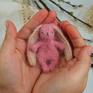 Op de afbeelding: Een klein roze gebreid konijn met crèmekleurige oren. Het konijn wordt in de handpalmen van twee handen gehouden.