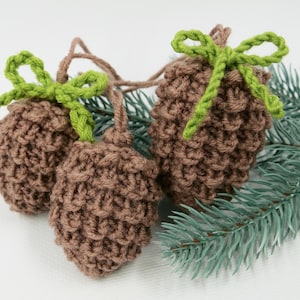 May include: Three brown crocheted pine cones with green yarn bows. The pine cones are sitting on a green pine branch.