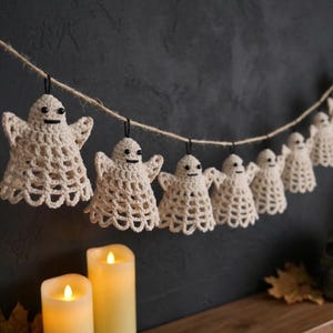 May include: A garland featuring a series of small, off-white crocheted ghost figures suspended from a length of natural jute twine. Each ghost has a bell-shaped body with an openwork pattern, small black bead eyes, and a stitched horizontal mouth.