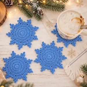 May include: Three blue crochet coasters shaped like snowflakes with intricate circular centers and pointed edges. The coasters are arranged on a light-colored surface alongside a ceramic mug filled with a beverage, pine branches, and ornaments.