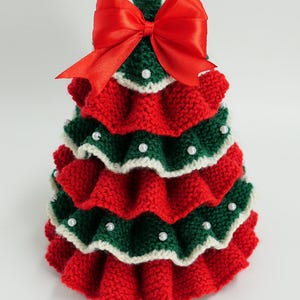 K&ouml;nnte beinhalten: Handgefertigte Weihnachtsbaumdekoration. Der Baum ist aus roter und gr&uuml;ner Wolle gestrickt, mit wei&szlig;em Rand und kleinen wei&szlig;en Perlen verziert. Oben befindet sich eine gro&szlig;e rote Satinschleife. Festliche Weihnachtsdekoration.