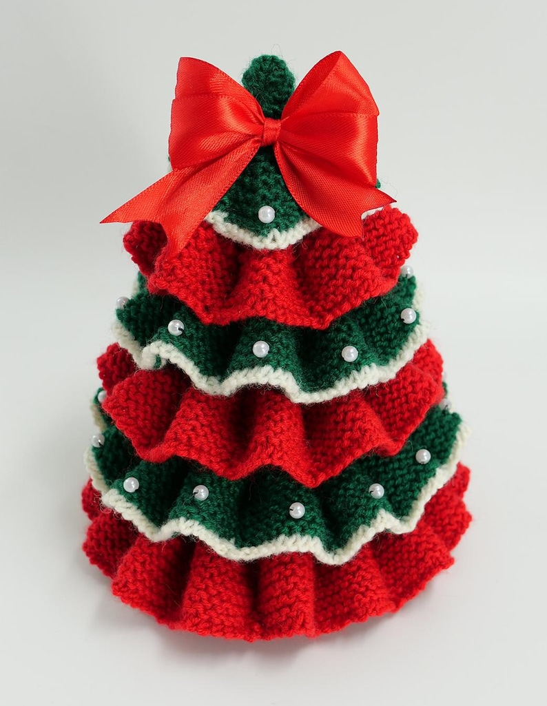K&ouml;nnte beinhalten: Handgefertigte Weihnachtsbaumdekoration. Der Baum ist aus roter und gr&uuml;ner Wolle gestrickt, mit wei&szlig;em Rand und kleinen wei&szlig;en Perlen verziert. Oben befindet sich eine gro&szlig;e rote Satinschleife. Festliche Weihnachtsdekoration.