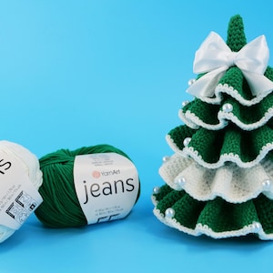 May include: A crocheted Christmas tree with white and green yarn, adorned with a white bow and pearl accents. Two balls of yarn, one white and one green, are labeled "jeans". The tree is on a blue background.