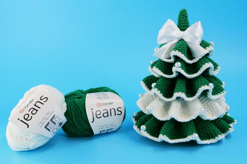 May include: A crocheted Christmas tree with white and green yarn, adorned with a white bow and pearl accents. Two balls of yarn, one white and one green, are labeled "jeans". The tree is on a blue background.